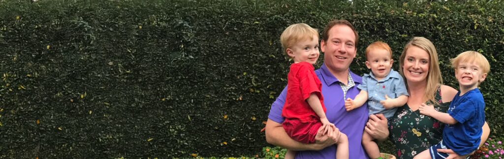 Jake Carris and his wife Erin smile as they hold their three young blonde boys, Rhett, Archer, and Calvin, standing in front of a dense green ivy wall.