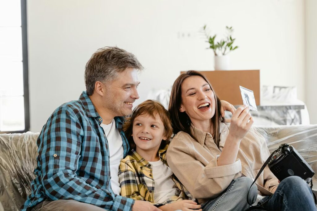 A smiling family of three sits on a couch, surrounded by moving boxes. The mother, on the right, holds up a small photo, laughing while looking at the image. The father, in a blue plaid shirt, and their young son smile happily beside her.