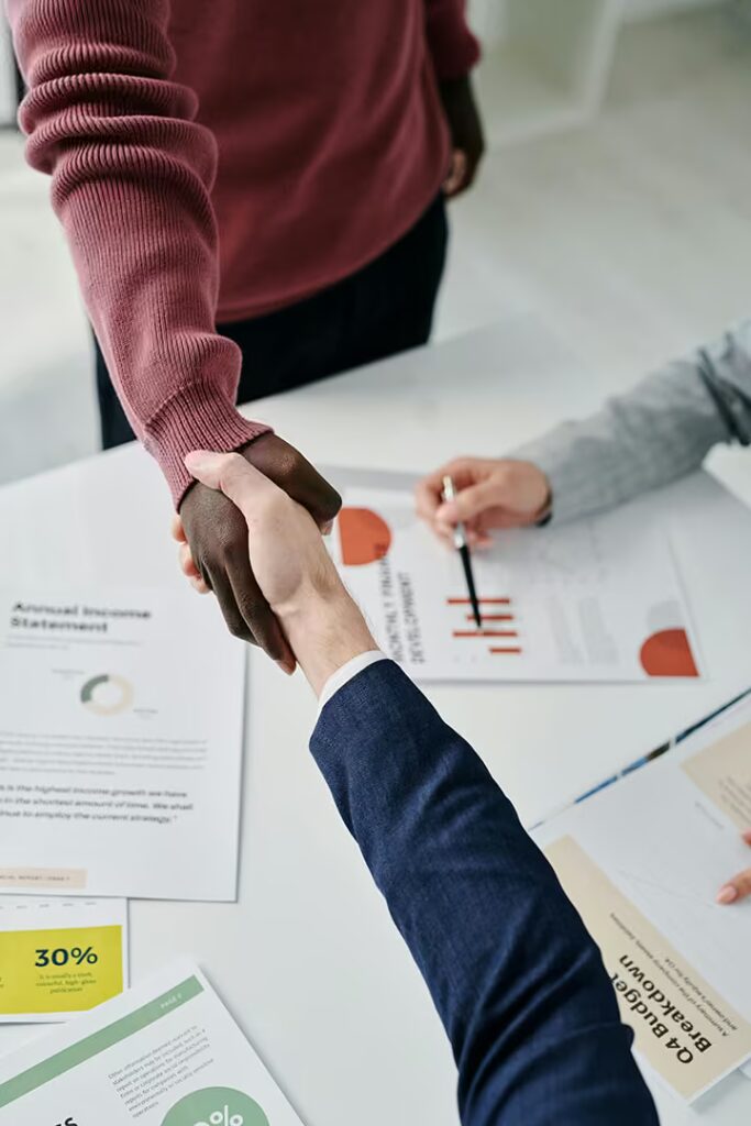 A close-up shot of two people shaking hands over a white conference table covered with various business documents, including a "Q4 Budget Breakdown" and "Annual Income Statement." One person is wearing a deep red sweater and the other is wearing a navy suit jacket.